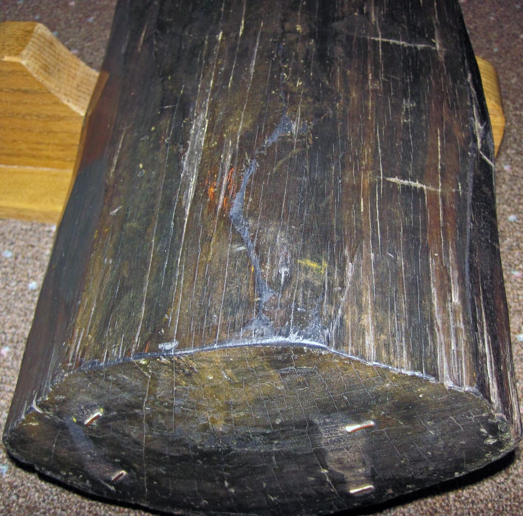 A weathered, dark hardwood specimen with visible splits and old nail fragments, displayed on a wooden stand