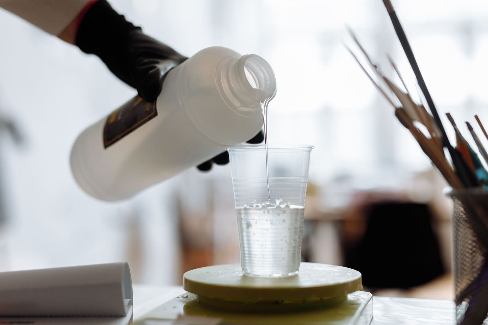 A gloved hand pouring clear liquid resin from a bottle into a plastic measuring cup