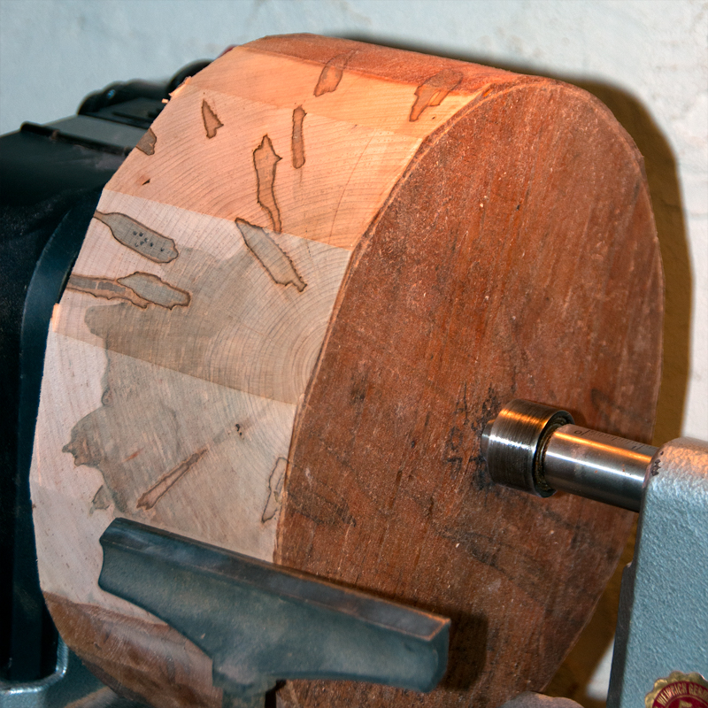 A rough blank of Ambrosia Maple mounted to a wood lathe before turning, the workshop behind